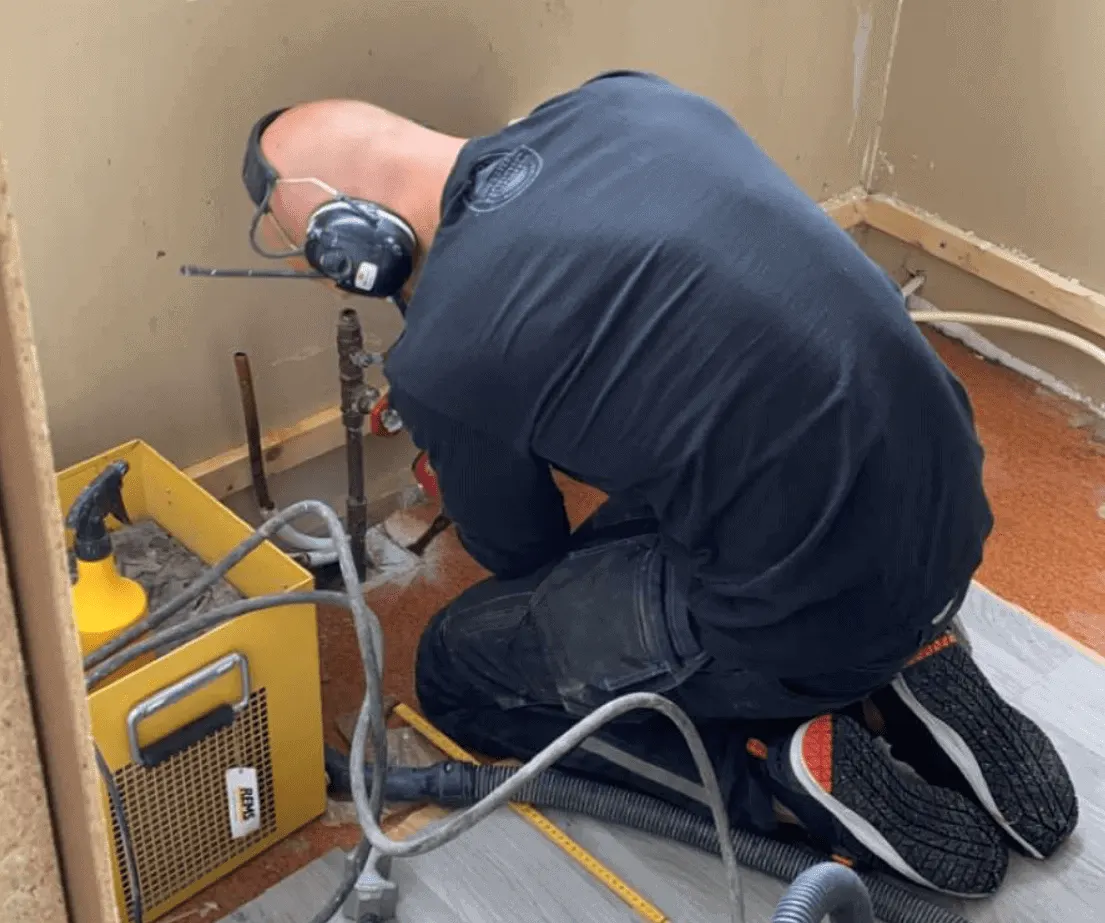 Rørlegger i arbeid ved et åpnet gulv med rør og trykktester.